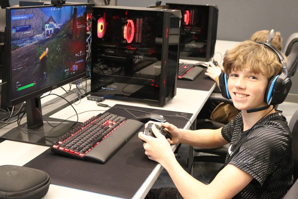 alt= headset smiles at the camera as he plays Fortnite on a gaming computer.