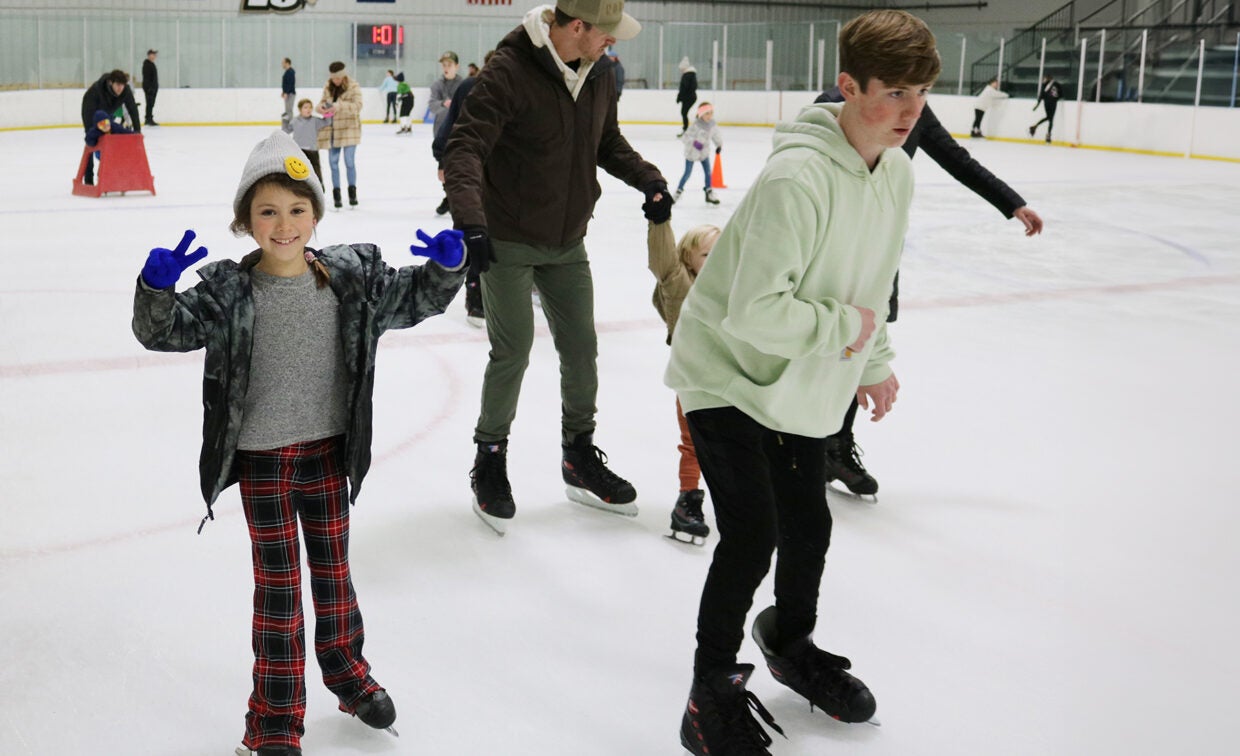 A girl in ice skates shows a peace symbol with both hands while she skates around the ice with other skaters.