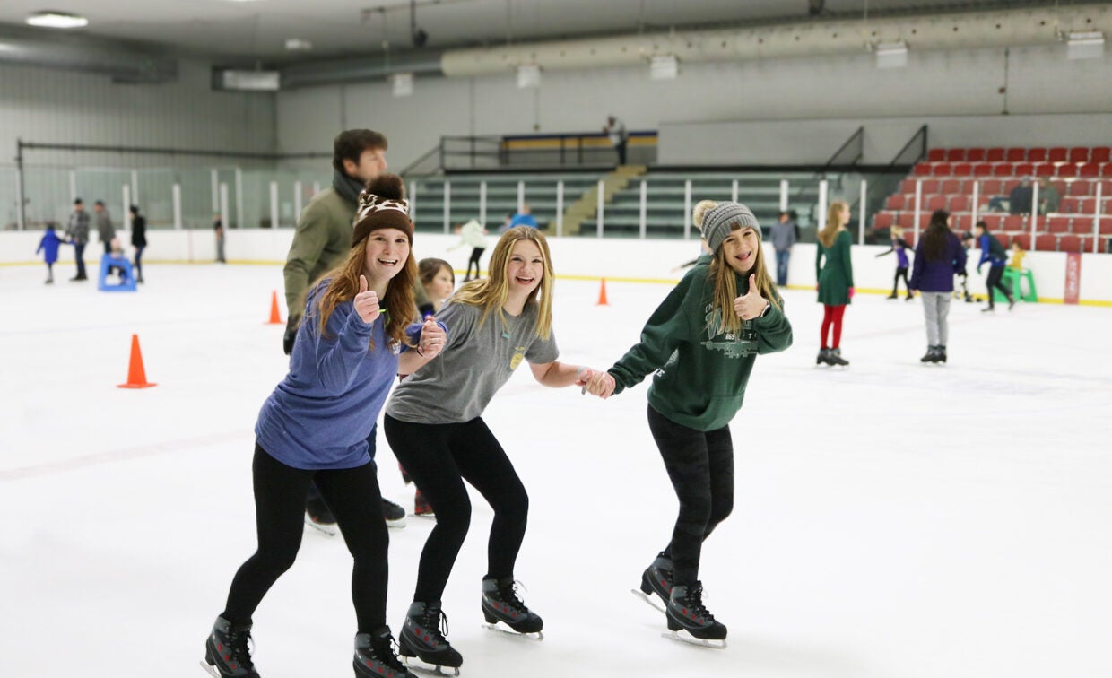 Three girls have fun skating around the ice with other skaters in the background.