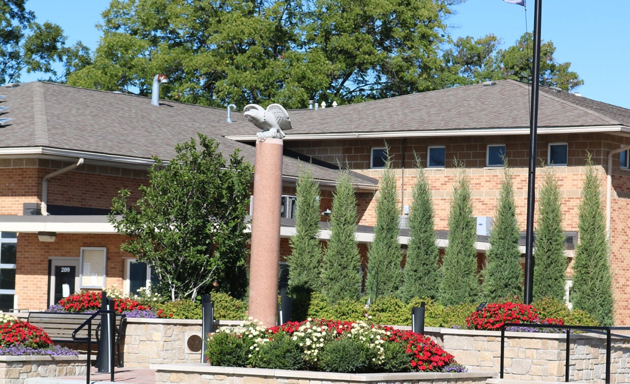 a Vietnam War memorial with a stone eagle sitting atop a stone pillar surrounded flower beds with eight fir trees and a bench.