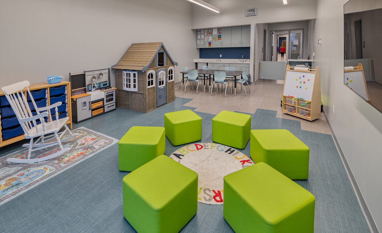 alt= Six green ottoman chairs are circled up in the Kids' Club room with a white rocking chair, play kitchen and house, a table with six chairs and a magnetic white board.