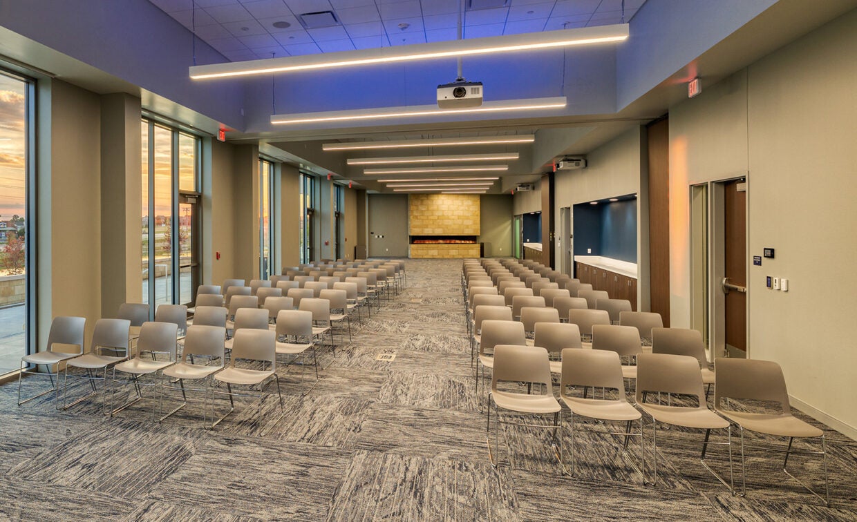 alt= Rows of chairs are set up in a large room with windows and a projector with a lit fireplace in the back of the room.