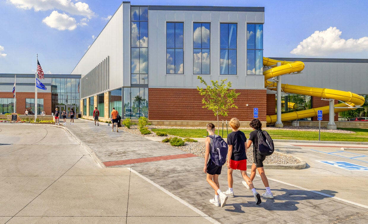 alt= Three boys walk up to the front of a building with a yellow slide on the outside and the American flag on a flag pole out front.