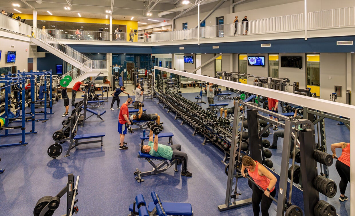 alt= Men and women work out in a fitness area that contains a lot of free weights and dumb bells.