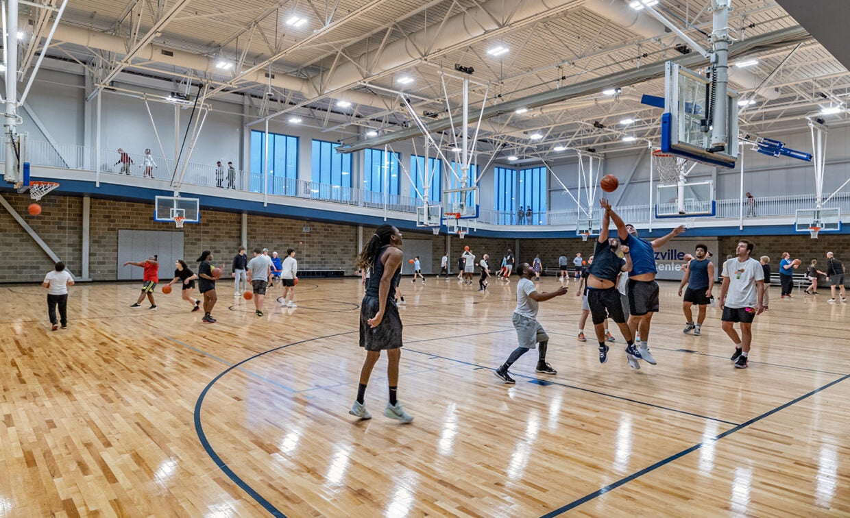 alt= Lots of men play basketball in a large gym while people walk around the elevated track.