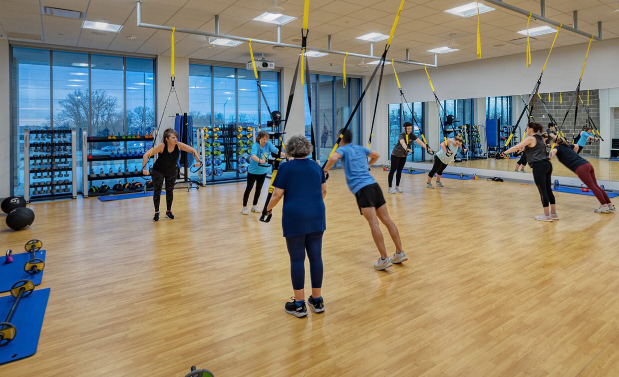 alt= Eight people lean on straps hung from the ceiling in a fitness class room,