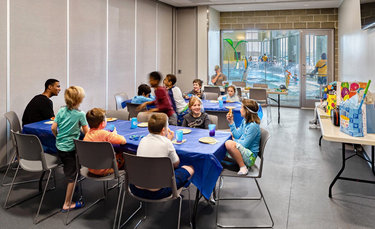 alt= Boys and girls celebrate a birthday in a room next to the recreation pool with tables and chairs for pizza and presents.