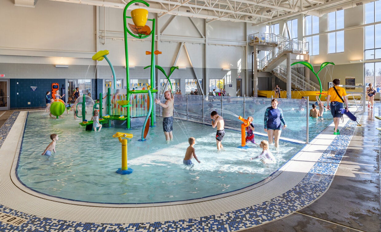 alt= People play in the water in front of the playground that is in the pool.