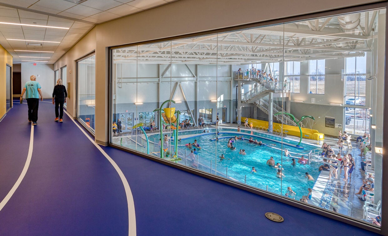 alt= A man and woman walk around an indoor track that overlooks the recreation pool with lots of swimmers.