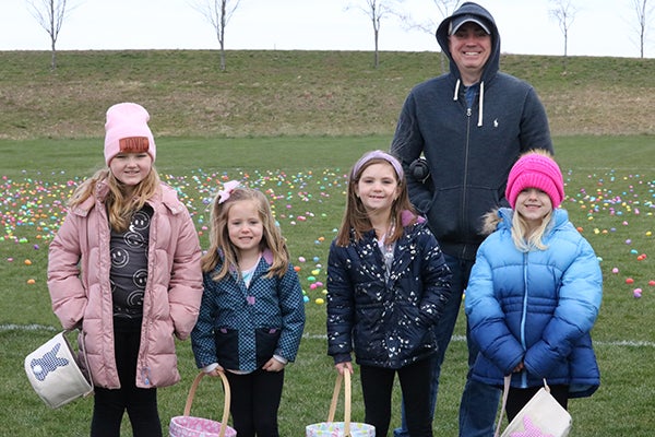 alt= Four girls in heavy coats stand on a field covered in Easter eggs holding baskets with a man in navy standing behind them.