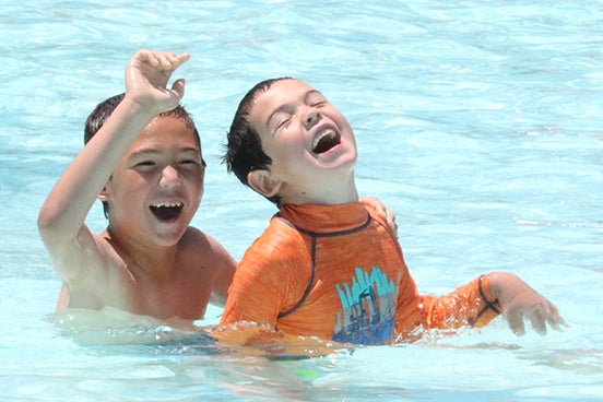 alt= Two boys, one wearing an orange, long-sleeve swim shirt, laugh and play in the pool.