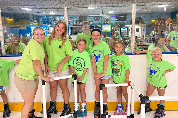 alt= Two female camp counselors in lime green T-shirts stand in roller skates with four girls also in lime green T-shirts on the roller skating rink with other campers looking on behind the plexiglass.