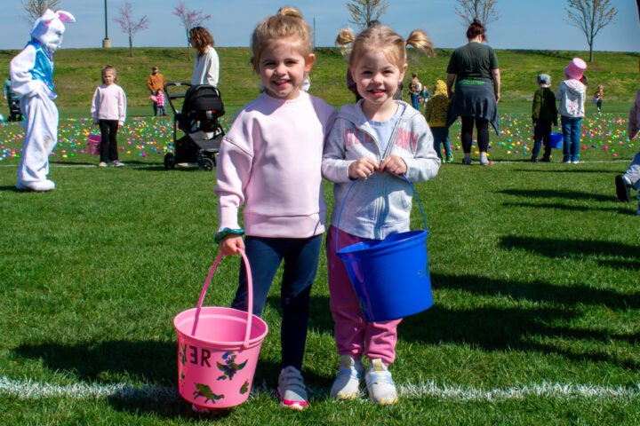 alt= two girls hold Easter baskets and smile and hug in front of a field with Easter eggs scattered in the grass