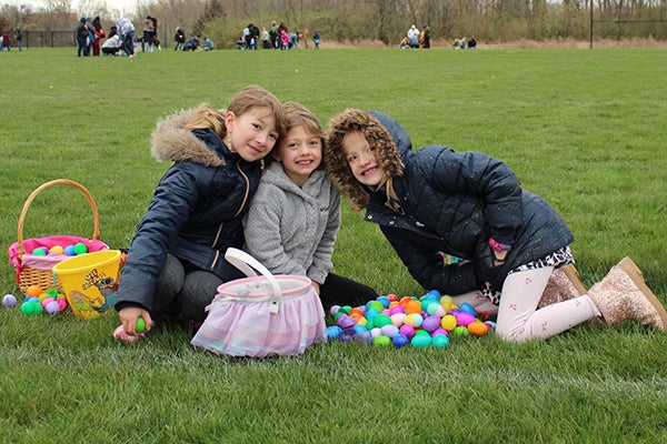 alt= three girls dressed in warm coats, kneel on the grass with a lot of plastic Easter eggs on the grass with them.