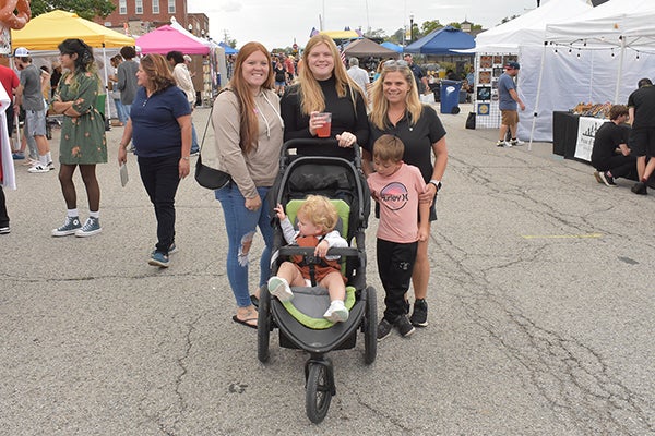 alt= Three women, a young boy and a baby in a stroller pose for a picture in the middle of the street lined with vendor booths.