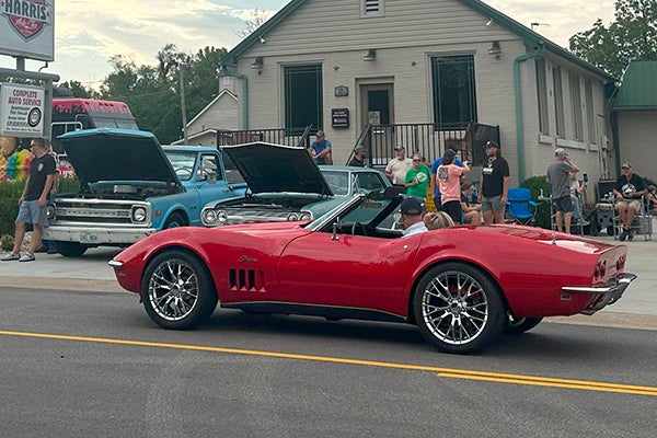 alt= An older red corvette drives down the street in front of two parked blue vehicles with their hoods raised.