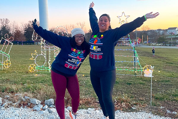 alt= Two women stand in front of a holiday light display and look happy with arms outstretched in nave sweatshirts that say may all your miles be bright, holiday night lights. It is still light outside.