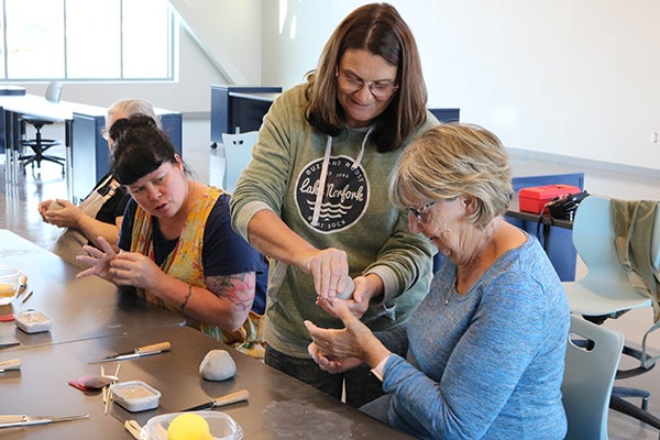 alt= A woman stands and teaches three other women how to make a pinch pot out of clay as they sit at a table with clay working toools.