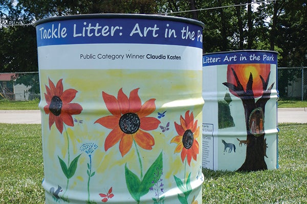 alt= Two trash cans that are colorfully wrapped. The front one has drawings of flowers and the back one has a tree. Both tops read Tackle Litter: Art in the Park.