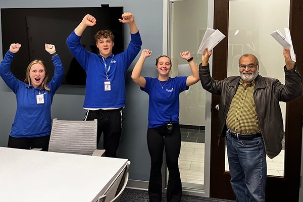 alt= Two women and one man in blue shirts raise their arms and cheer on another man holding up several envelopes.