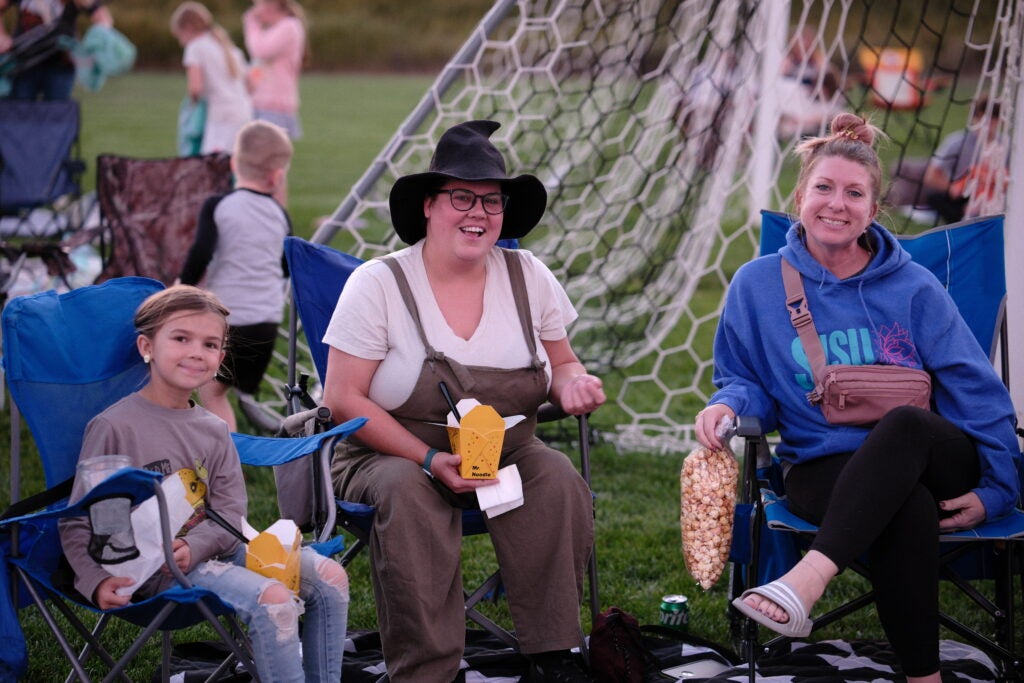 three girls sitting in lawn chairs on a soccer field eating snacks