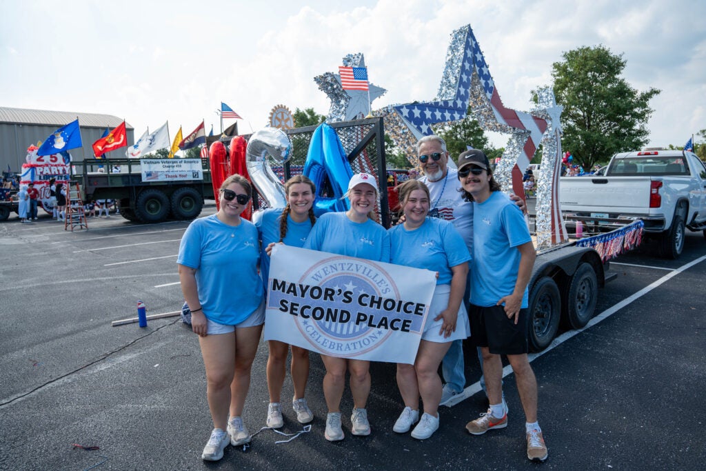 Group of people holding a banner, in front of their parade float with the mayor