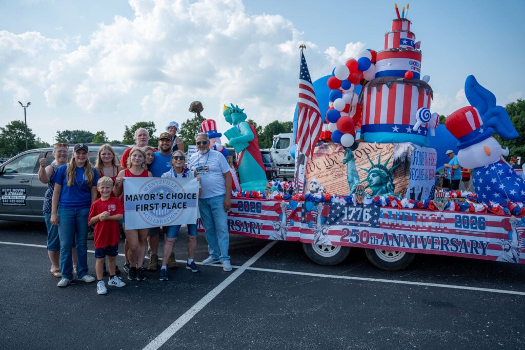 Group of people holding a banner, in front of their parade float with the mayor